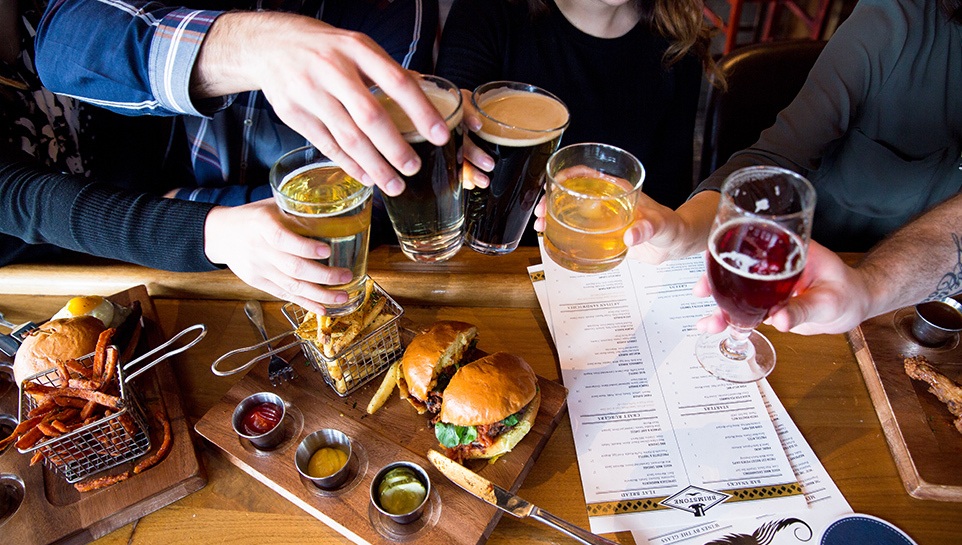 Hands toasting beers at Brimstone with a bar full of food